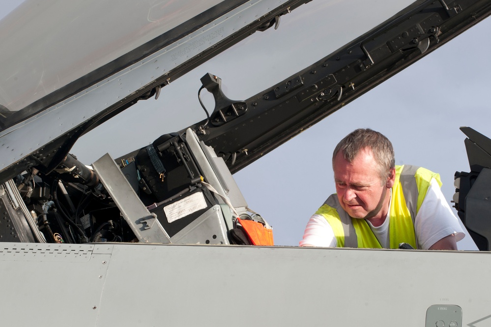 A ground crew technician wearing a high-visibility yellow vest is inspecting or servicing the open cockpit of a military jet aircraft. The canopy is raised, exposing the cockpit interior including avionics equipment, wiring, and instrumentation. The work appears to be a pre-flight or maintenance check conducted on an airfield.