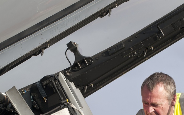 A ground crew technician wearing a high-visibility yellow vest is inspecting or servicing the open cockpit of a military jet aircraft. The canopy is raised, exposing the cockpit interior including avionics equipment, wiring, and instrumentation. The work appears to be a pre-flight or maintenance check conducted on an airfield.