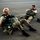 Two military personnel in full tactical gear are seated on what appears to be a tarmac or airfield surface. Both individuals are wearing camouflage uniforms and are equipped with parachute rigs and other tactical equipment. They appear to be resting or waiting, possibly before or after a jump operation.