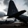 The image shows U.S. Air Force C-130 Hercules transport aircraft on a flight line at dusk or dawn under a dramatic cloudy sky. The foreground aircraft displays tail code 'FT' and serial number 'AF 65 986', with personnel gathered beneath the rear of the aircraft. Multiple C-130s are visible on the tarmac, suggesting a forward operating base or deployment location.