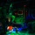 Military personnel are inside what appears to be a large military transport aircraft, likely a CH-47 Chinook or C-130, during a nighttime operation. The interior is illuminated by multicolored lights including green, red, and blue, casting dramatic shadows on the soldiers and equipment. Personnel appear to be preparing for or conducting a parachute or airborne insertion operation.
