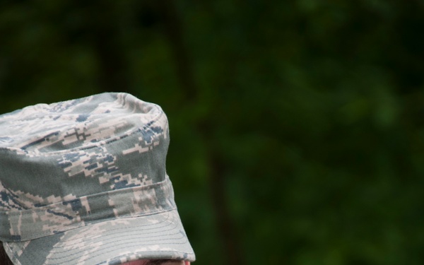 A U.S. Air Force servicemember in Airman Battle Uniform (ABU) is intently reading a military lensatic compass held close to eye level. The individual appears to be conducting land navigation training or a field exercise in an outdoor, wooded environment. The uniform text 'U.S. AIR' is clearly visible on the chest.