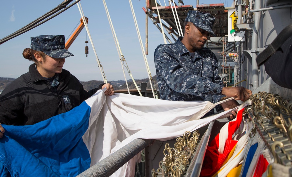 Two U.S. Navy sailors in Navy Working Uniform (NWU) digital camouflage are handling signal flags aboard a naval vessel. They appear to be preparing or organizing nautical signal flags, which are stored on a flag bag rail with brass snap hooks. The setting is the deck of a military ship with rigging, masts, and superstructure visible in the background.