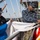 Two U.S. Navy sailors in Navy Working Uniform (NWU) digital camouflage are handling signal flags aboard a naval vessel. They appear to be preparing or organizing nautical signal flags, which are stored on a flag bag rail with brass snap hooks. The setting is the deck of a military ship with rigging, masts, and superstructure visible in the background.
