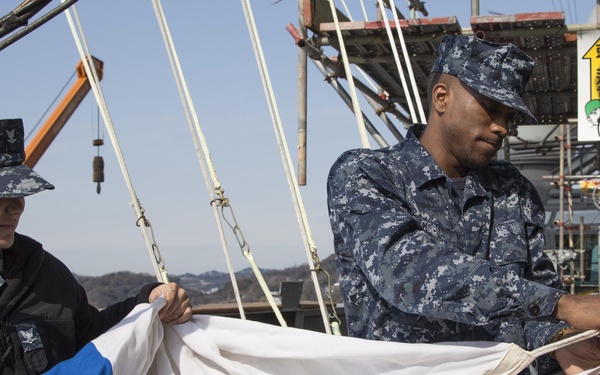 Two U.S. Navy sailors in Navy Working Uniform (NWU) digital camouflage are handling signal flags aboard a naval vessel. They appear to be preparing or organizing nautical signal flags, which are stored on a flag bag rail with brass snap hooks. The setting is the deck of a military ship with rigging, masts, and superstructure visible in the background.