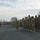A formation of U.S. Army soldiers in camouflage uniforms (ACU/OCP pattern) stands at attention in a parking lot during what appears to be a military ceremony, likely a deployment sendoff or homecoming event. A crowd of civilians lines the left side, many holding American flags and signs. The setting appears to be a suburban or small-town location during late fall or winter, based on the bare trees and overcast sky.