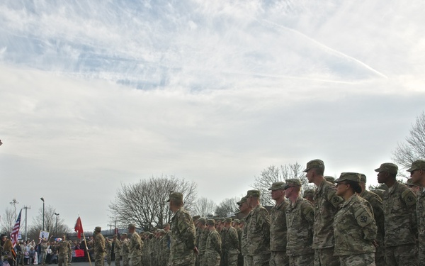 A formation of U.S. Army soldiers in camouflage uniforms (ACU/OCP pattern) stands at attention in a parking lot during what appears to be a military ceremony, likely a deployment sendoff or homecoming event. A crowd of civilians lines the left side, many holding American flags and signs. The setting appears to be a suburban or small-town location during late fall or winter, based on the bare trees and overcast sky.