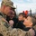 A U.S. Army soldier in camouflage uniform is being greeted by what appears to be his family, including a woman and two children, in an emotional reunion. The soldier is embracing a boy while a girl wearing red Minnie Mouse ears smiles nearby. The setting appears to be a military installation or base, suggesting a homecoming event.