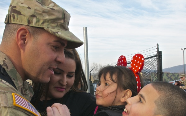 A U.S. Army soldier in camouflage uniform is being greeted by what appears to be his family, including a woman and two children, in an emotional reunion. The soldier is embracing a boy while a girl wearing red Minnie Mouse ears smiles nearby. The setting appears to be a military installation or base, suggesting a homecoming event.