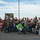 A large group of family and friends has gathered outdoors in a parking lot to welcome home a U.S. Army soldier named Elias. The group is holding a Puerto Rican flag and a black banner reading 'Welcome Home Elias! We love you!' with the U.S. Army logo. The crowd includes adults, children, and infants, with several people wearing matching black U.S. Army-themed shirts.