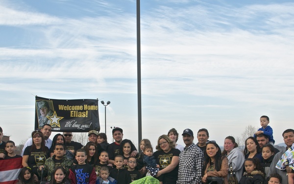 A large group of family and friends has gathered outdoors in a parking lot to welcome home a U.S. Army soldier named Elias. The group is holding a Puerto Rican flag and a black banner reading 'Welcome Home Elias! We love you!' with the U.S. Army logo. The crowd includes adults, children, and infants, with several people wearing matching black U.S. Army-themed shirts.