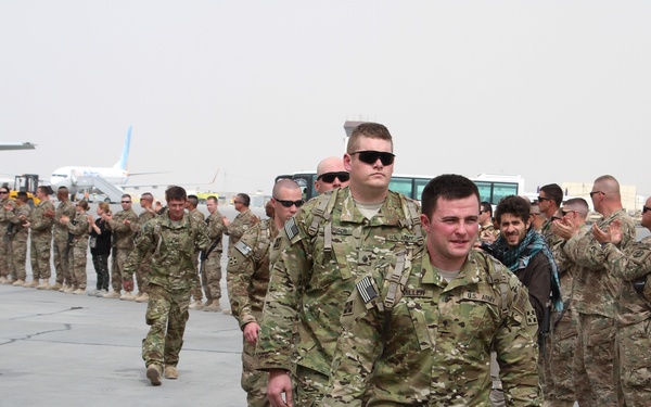 A group of U.S. Army soldiers in MultiCam/OCP camouflage uniforms walk through a corridor of applauding fellow service members on what appears to be a military airfield tarmac. The scene depicts a traditional 'clap-out' or welcome ceremony, likely marking a deployment departure or return. Aircraft and military infrastructure are visible in the background.
