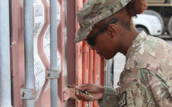 A U.S. Army soldier in MultiCam/OCP camouflage uniform is working with a lock on a shipping container. The soldier is wearing sunglasses and a patrol cap, and appears to be either locking or unlocking a padlock on the container's locking bar mechanism. A military vehicle is visible in the background.
