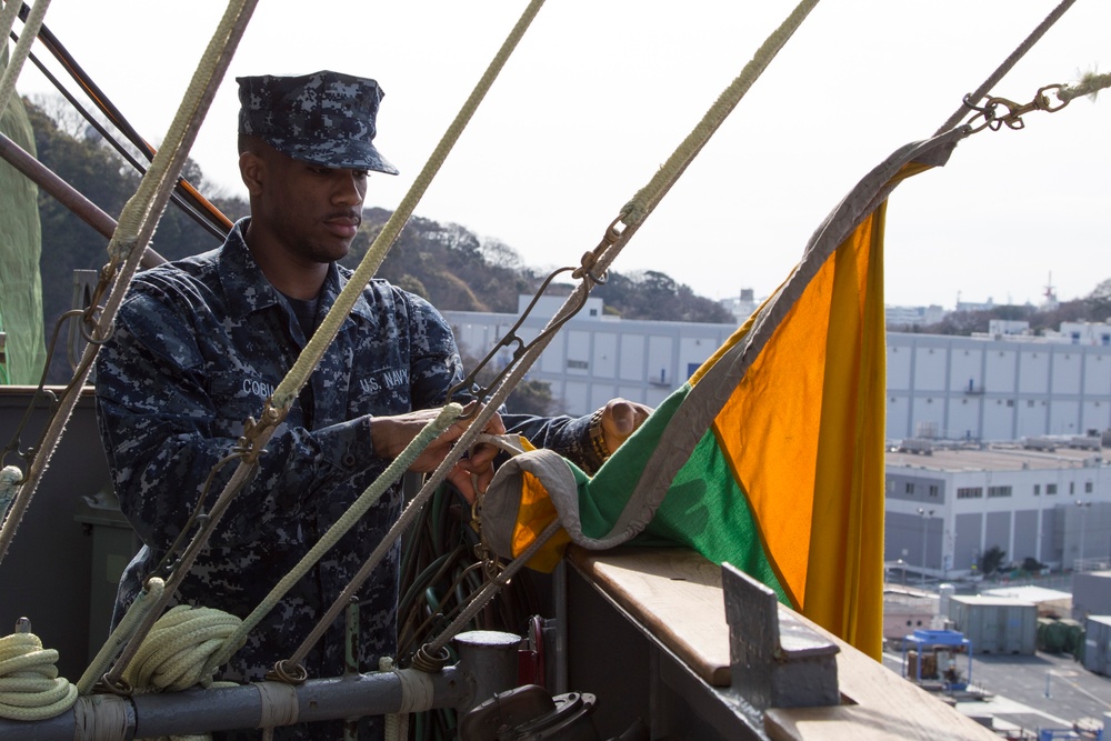 A U.S. Navy sailor in digital camouflage (NWU Type I) uniform is working with signal flags or rigging on what appears to be the elevated deck or mast area of a naval vessel. The sailor's name tape reads 'COBI' and the branch tape reads 'U.S. NAVY'. The background shows a port or naval base facility with industrial buildings and hillside vegetation, suggesting a location such as Yokosuka Naval Base, Japan.