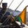 A U.S. Navy sailor in digital camouflage (NWU Type I) uniform is working with signal flags or rigging on what appears to be the elevated deck or mast area of a naval vessel. The sailor's name tape reads 'COBI' and the branch tape reads 'U.S. NAVY'. The background shows a port or naval base facility with industrial buildings and hillside vegetation, suggesting a location such as Yokosuka Naval Base, Japan.