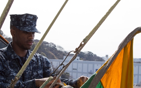A U.S. Navy sailor in digital camouflage (NWU Type I) uniform is working with signal flags or rigging on what appears to be the elevated deck or mast area of a naval vessel. The sailor's name tape reads 'COBI' and the branch tape reads 'U.S. NAVY'. The background shows a port or naval base facility with industrial buildings and hillside vegetation, suggesting a location such as Yokosuka Naval Base, Japan.