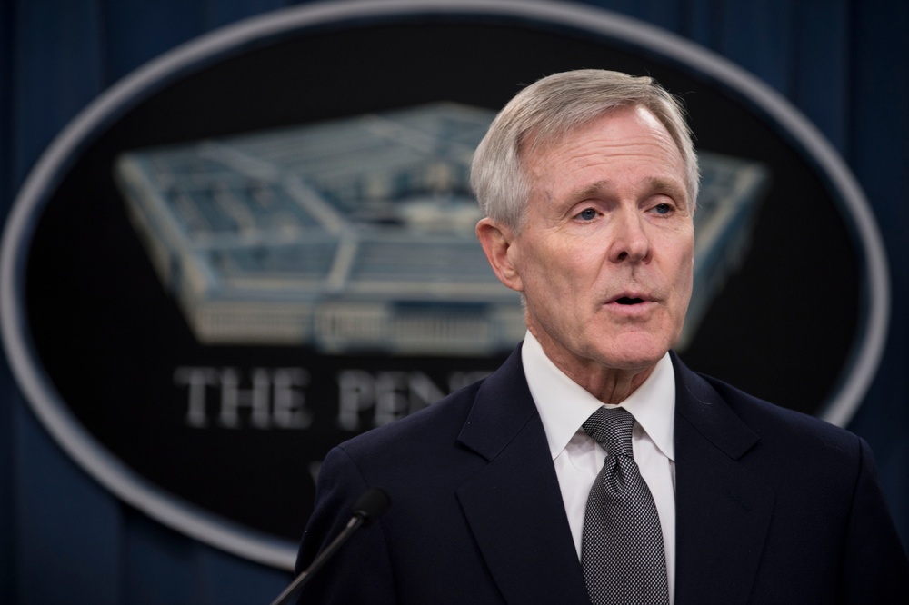A man in a dark suit and gray tie is speaking at a podium in front of the Pentagon seal. The setting appears to be an official press briefing room at the Pentagon. The subject appears to be a senior official delivering remarks or answering questions.