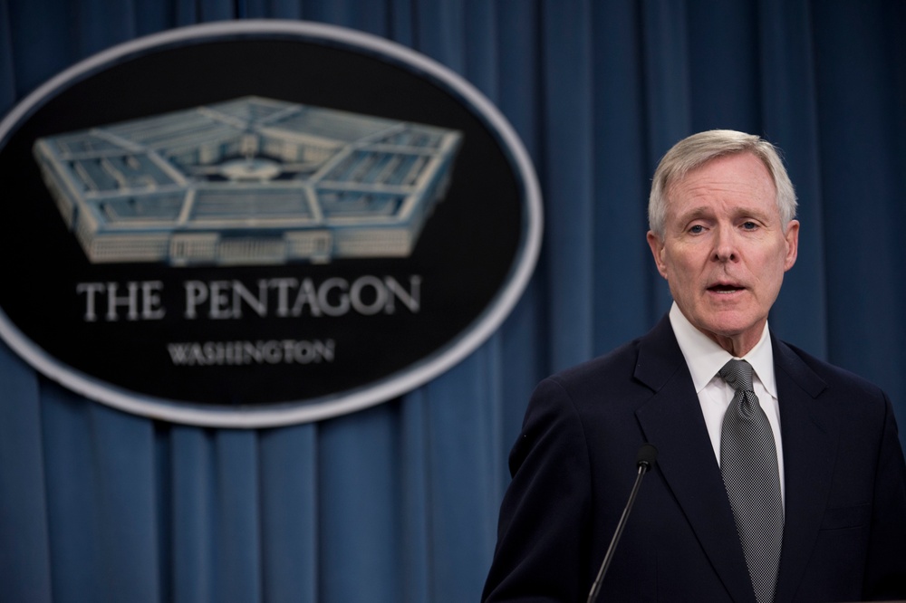 A man in a dark suit is speaking at a podium with microphones at The Pentagon in Washington. The official Pentagon seal is prominently displayed on a blue curtain backdrop behind him. The setting appears to be a formal press briefing or official announcement.