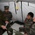 Chaplain exchange promotes brotherhood during MEFEX 2014 A military briefing or demonstration is taking place inside a field tent, where a Western military personnel (likely U.S. Marine based on MARPAT camouflage uniform) is showing equipment or medical supplies to several Asian military personnel. The individuals are gathered around a table with military gear, bags, and supplies laid out. The setting appears to be a joint military exercise or training event.