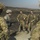 A group of military personnel from at least two different nations are gathered in an outdoor field setting, appearing to be in a briefing or debrief situation. One soldier wearing US Army ACU (Army Combat Uniform) with a visible American flag patch is addressing a group that includes soldiers in British MTP (Multi-Terrain Pattern) camouflage, some wearing ghillie suit headgear. The scorched ground in the background suggests a recent training exercise or controlled burn area.