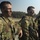 Three military personnel in camouflage uniforms are standing in an outdoor field environment, appearing to have recently completed a training exercise. They have camouflage face paint applied and show signs of physical exertion, with wet or sweaty appearances. The soldiers are carrying weapons and field equipment, with a forested area and open terrain visible in the background.