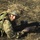 A soldier in prone firing position aims a rifle toward the camera on open ground. The individual is wearing British MTP (Multi-Terrain Pattern) camouflage uniform with a helmet adorned with natural vegetation (grass/straw) for concealment. The image appears to be from a military training exercise.