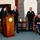 A formal military ceremony taking place in an ornate room, likely a government building. A civilian woman is speaking at a podium bearing an official seal, flanked by three U.S. Army soldiers in dress uniforms. A framed portrait of a man is displayed to the left on an easel, suggesting this may be a memorial or award ceremony.