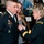 A military promotion or award ceremony is taking place indoors. A female Army officer is pinning a medal or insignia onto the uniform of Staff Sergeant Dimond, while a young woman stands closely behind him observing. The setting appears to be an official military venue with a portrait painting and other uniformed personnel visible in the background.