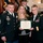 A formal U.S. Army Purple Heart award ceremony is taking place indoors. A soldier identified by nameplate as 'DIMOND' wearing dress uniform with a Purple Heart medal pinned to his chest stands alongside a civilian woman and a female Army officer, all holding a Purple Heart certificate. Additional uniformed soldiers are visible in the background near an American flag.