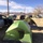 A campground scene in an arid, desert-like setting with mountains visible in the background. Multiple tents, vehicles, and RVs are spread across dry, grassy terrain under a partly cloudy blue sky. The foreground features a prominently branded green Coleman dome tent with its door open.