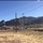 A desert landscape scene featuring a commercial or civic building complex with a parking area filled with vehicles and people, set against a dramatic rocky mountain range backdrop. The foreground consists of dry, sparse desert terrain with utility poles and power lines. The setting appears to be a southwestern U.S. location, likely New Mexico or Arizona, based on the terrain and mountain profile.
