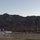 The image shows an outdoor sports facility, likely a soccer field, set against a dramatic rocky mountain backdrop in what appears to be a desert southwestern US location. Promotional banner flags in purple and blue are visible, along with a white metal soccer goal in the foreground. The scene appears to be captured during early morning or late afternoon based on the soft, low-angle lighting.