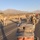 A selfie taken by a soldier in U.S. Army ACU (Army Combat Uniform) during what appears to be a ruck march or military road march event. Multiple soldiers carrying rucksacks are visible marching along a paved road in a desert environment with mountains in the background. A red guidon or flag is visible in the distance among the formation.