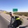 A U.S. Army soldier in ACU (Army Combat Uniform) is walking along the shoulder of a highway in a desert environment, carrying a large military rucksack with a high-visibility yellow strap. A green highway directional sign is visible in the background indicating US Route 70 West toward Las Cruces and US Route 70 East toward Alamogordo, placing the location in New Mexico near White Sands/Fort Bliss area. The terrain is arid with sparse desert vegetation and mountains visible in the distance.