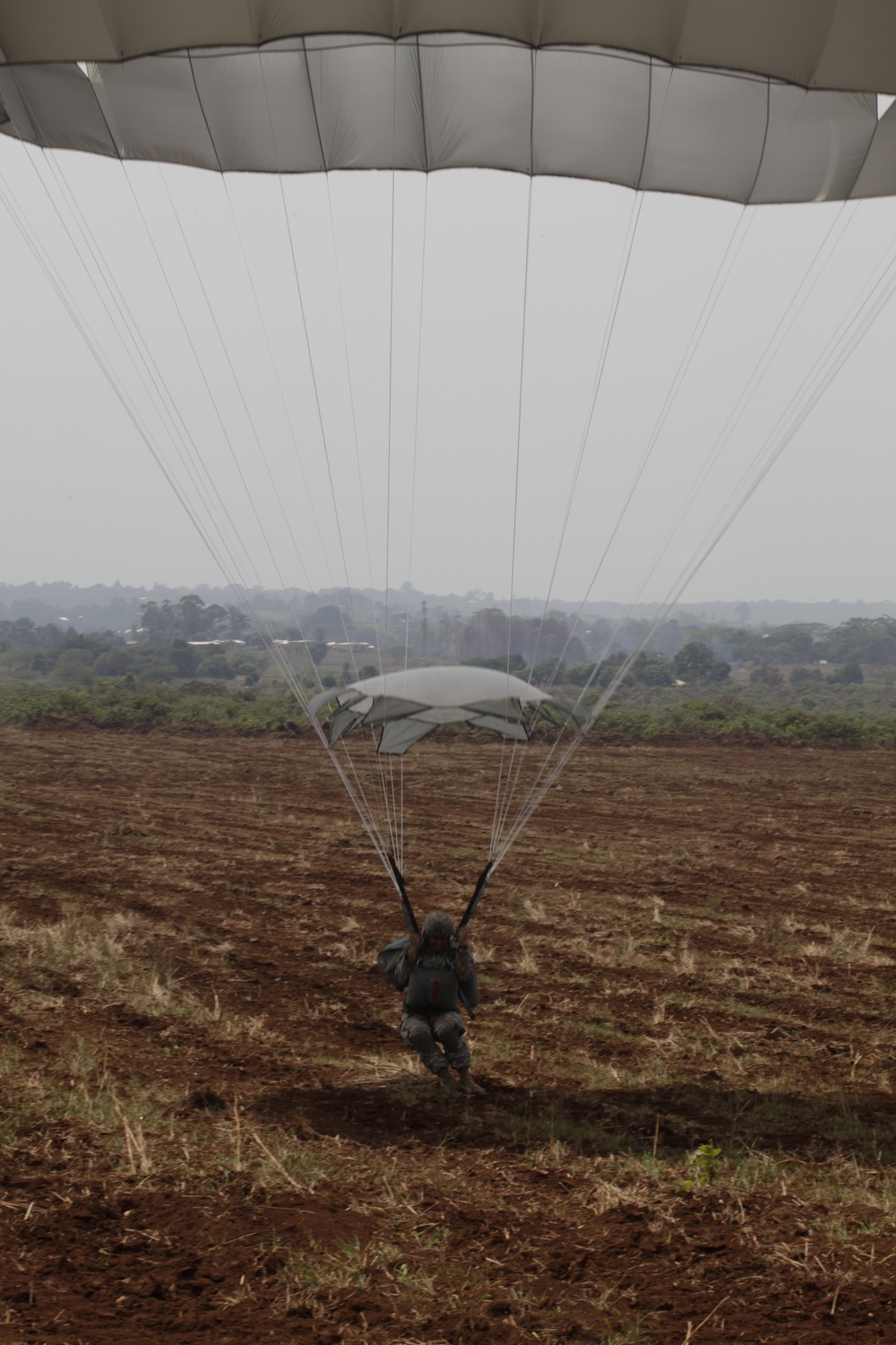 A military paratrooper is captured at the moment of landing on a large open dirt field, with a white/gray parachute canopy fully deployed overhead. A second parachute is visible in the mid-ground, suggesting a group jump exercise. The background shows a flat landscape with scattered trees and hazy overcast skies.