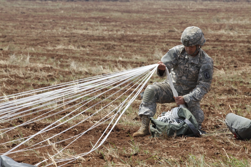 A U.S. Army soldier in ACU (Army Combat Uniform) is kneeling on a dirt field gathering and recovering parachute suspension lines after a jump. The soldier is wearing a combat helmet and knee pads, and is managing multiple white parachute cords that fan out across the ground. A parachute bag and additional gear are visible nearby on the reddish-brown soil.