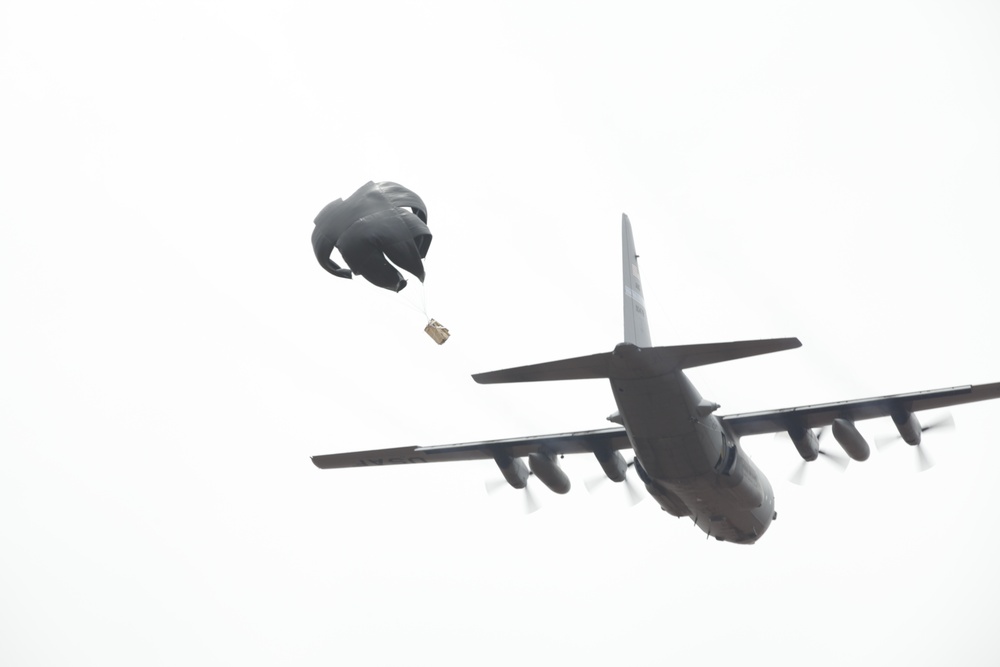 A military transport aircraft, consistent with a C-130 Hercules, is captured in flight performing an aerial cargo drop. A parachute has deployed and is carrying a small cargo bundle away from the aircraft. The image is taken from a low angle against an overcast, washed-out sky.