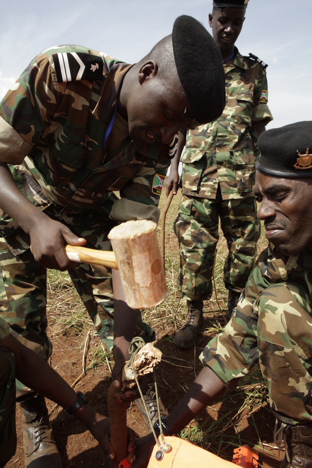 Several military personnel in camouflage uniforms are engaged in a field activity involving driving a stake or peg into the ground using a wooden mallet. One soldier is actively swinging the mallet while another crouches to guide the stake. A third soldier stands observing in the background.