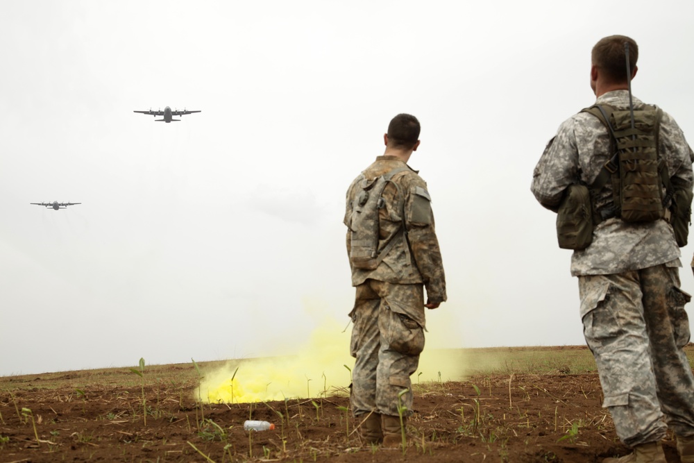 Two soldiers in US Army ACU (Army Combat Uniform) camouflage are observing military aircraft flying overhead in an overcast sky. A yellow smoke grenade has been deployed on the ground in front of them, likely used as a visual marker or signal for the aircraft. This appears to be a military training exercise or operational coordination activity involving ground troops and air assets.