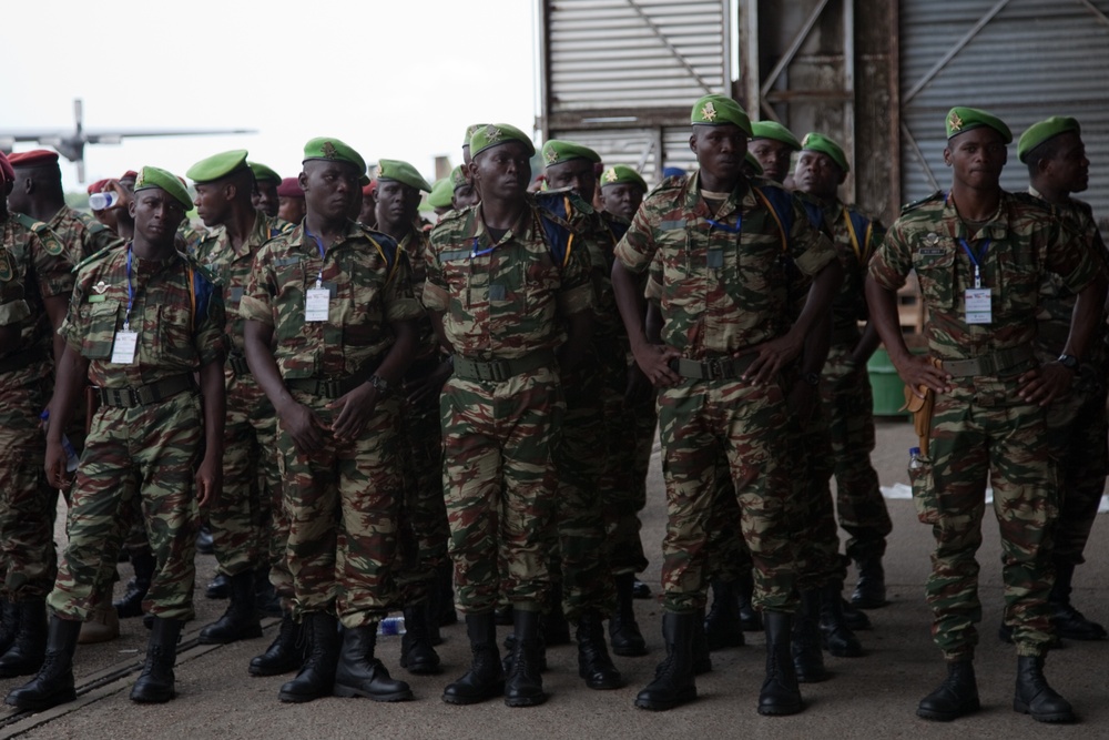 A large group of military personnel standing in formation, wearing matching camouflage uniforms and green berets. They appear to be assembled at what looks like a warehouse or hangar facility. Most personnel are wearing identification badges/lanyards, suggesting an organized official event or deployment.