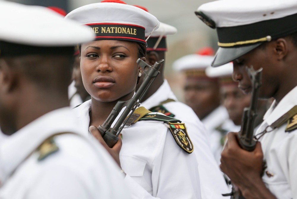 The image shows military personnel in white naval dress uniforms participating in what appears to be a formal parade or ceremony. A female sailor is prominently featured in the foreground, wearing a cap inscribed with 'MARINE NATIONALE' and carrying a rifle at shoulder arms. The shoulder patch and uniform details suggest this is likely a Francophone African navy, possibly Senegalese or Cameroonian.