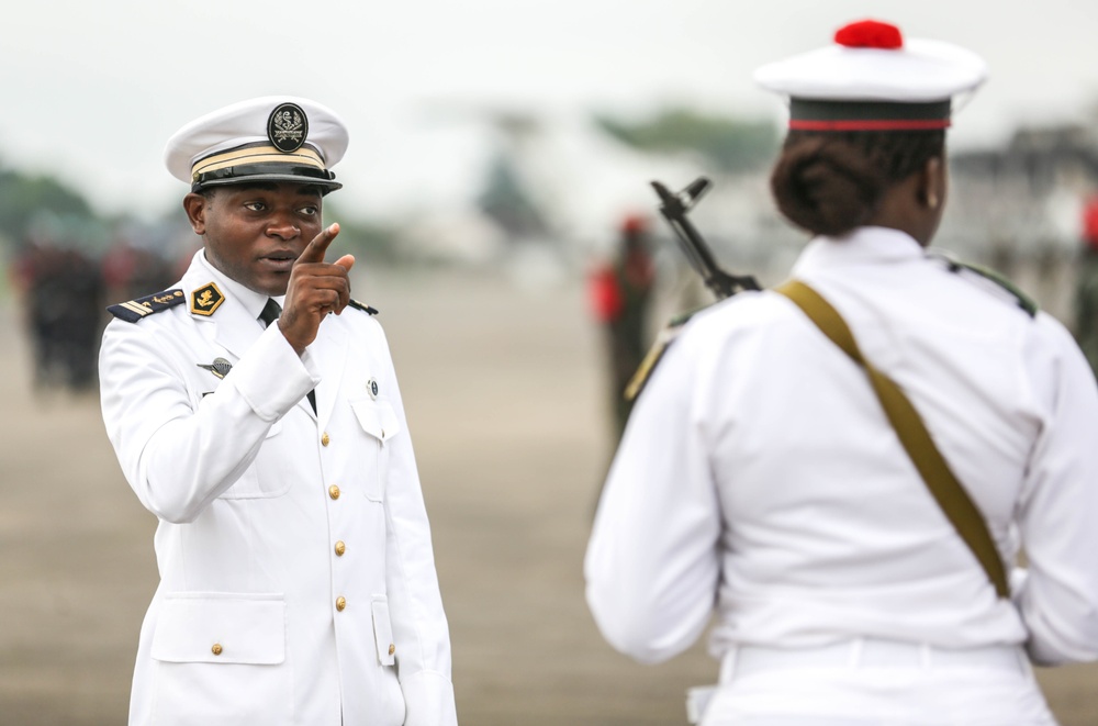 A military parade or ceremonial event is taking place on an open tarmac or parade ground. A senior officer in a white naval dress uniform is giving instructions or commands, pointing toward a soldier in white ceremonial uniform carrying a rifle. The setting appears to be an official military ceremony with multiple personnel visible in the background.