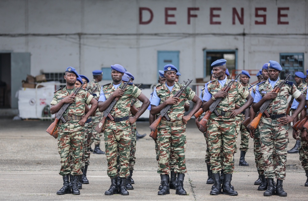 A formation of military soldiers standing at attention in front of a building marked 'DEFENSE'. The soldiers are wearing camouflage uniforms with blue berets and carrying rifles, suggesting a formal military parade or ceremonial drill. The setting appears to be a military installation or airbase.