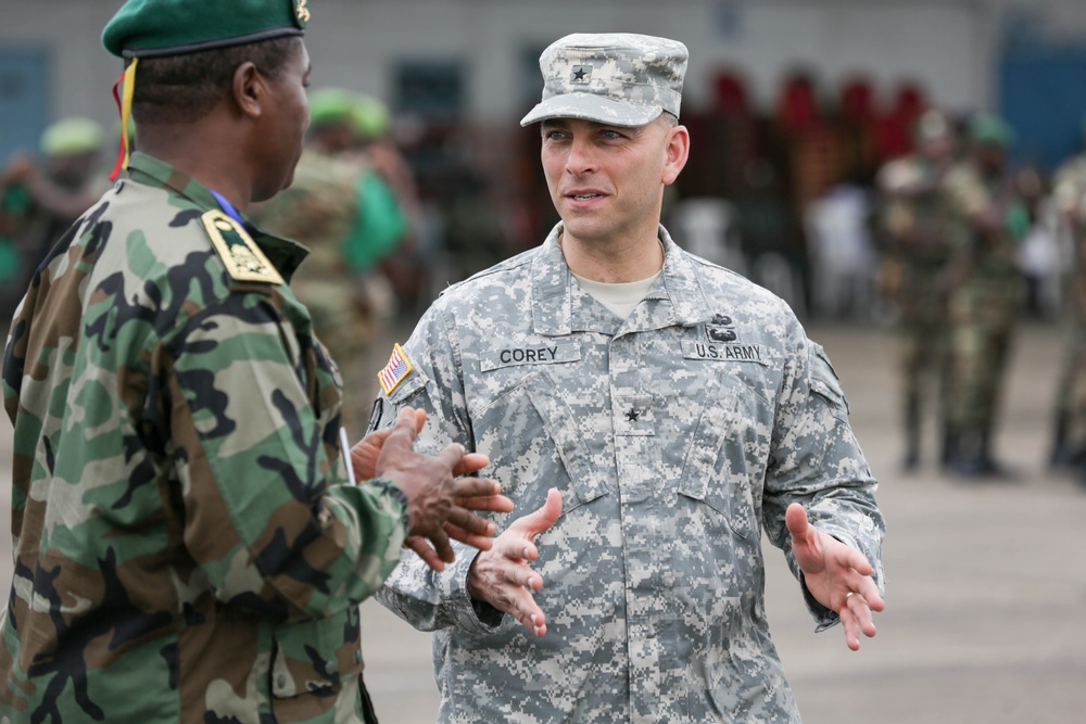 A U.S. Army officer wearing ACU (Army Combat Uniform) with the name tape 'COREY' and a single star insignia indicating the rank of Brigadier General is engaged in conversation with a foreign military officer in woodland camouflage. The setting appears to be an outdoor military event or ceremony, with additional military personnel visible in the background. The interaction suggests a bilateral or multinational military engagement.