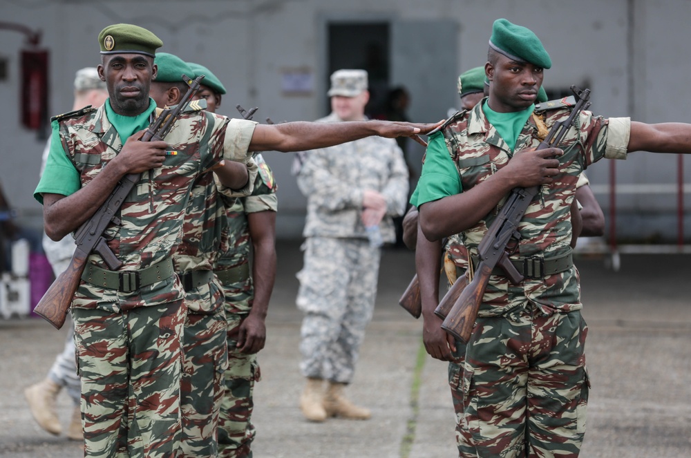 The image shows military personnel in formation, likely during a drill or ceremonial parade. Two foreground soldiers wearing green berets and camouflage uniforms carry rifles at port arms, with one extending an arm to maintain spacing. In the background, additional soldiers and what appears to be a US military personnel in ACU pattern uniform are visible.