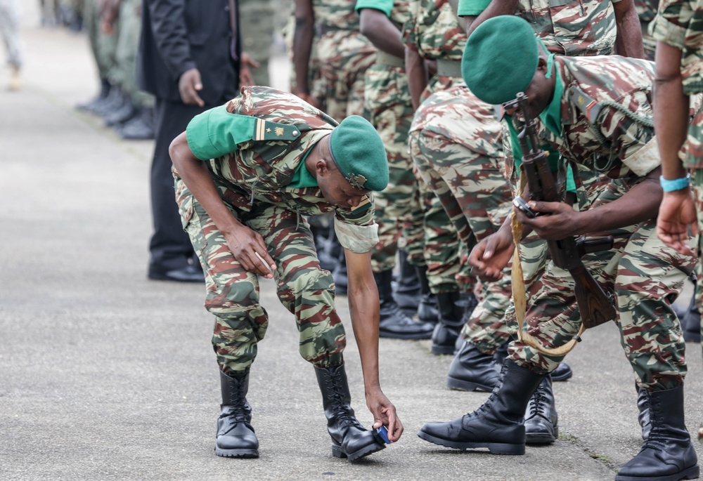 The image shows a group of military personnel in camouflage uniforms and green berets, apparently preparing for or participating in a formal parade or inspection. One soldier in the foreground is bent down adjusting something at the boot level of another soldier, possibly fixing a boot or placing a marker on the ground. The soldiers are equipped with rifles and wear matching green-and-brown camouflage uniforms with black combat boots.