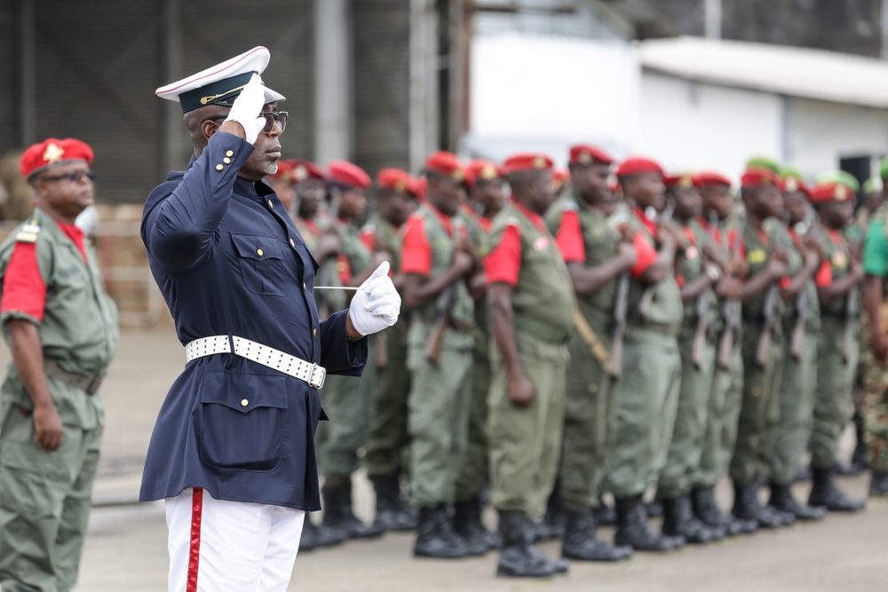 A military parade or ceremonial formation is taking place outdoors. A senior officer in a formal navy blue dress uniform with white gloves and cap is saluting while holding a baton, standing before rows of soldiers in olive green fatigues and red berets. The scene appears to be an official military ceremony, likely in a sub-Saharan African country.