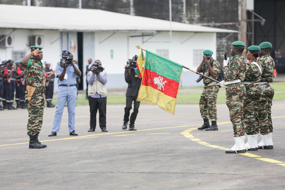 A military ceremony is taking place on a parade ground, featuring soldiers in camouflage uniforms and green berets. A senior officer on the left is rendering a salute toward a color guard bearing a tricolor flag (green, yellow, red) with insignia. Media personnel with cameras are documenting the event in the background.