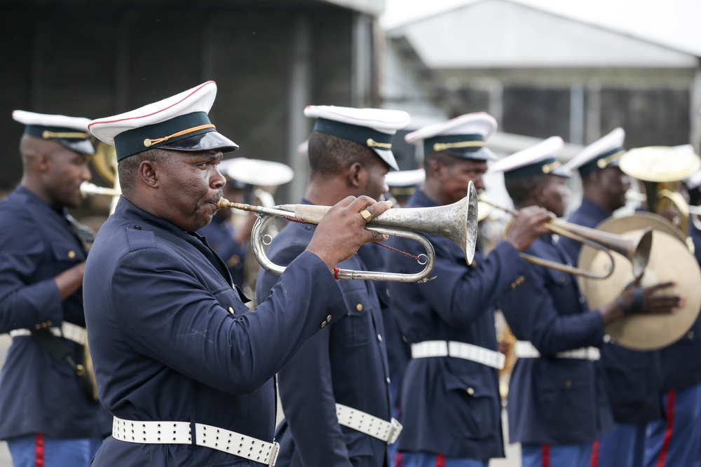 A military or ceremonial brass band is performing outdoors, with multiple musicians dressed in matching dark navy uniforms with white caps and white belts. The foreground features a musician playing a silver bugle or trumpet, while others behind him play similar brass instruments including what appears to be a sousaphone or tuba on the right. The setting appears to be an outdoor parade or official ceremony.