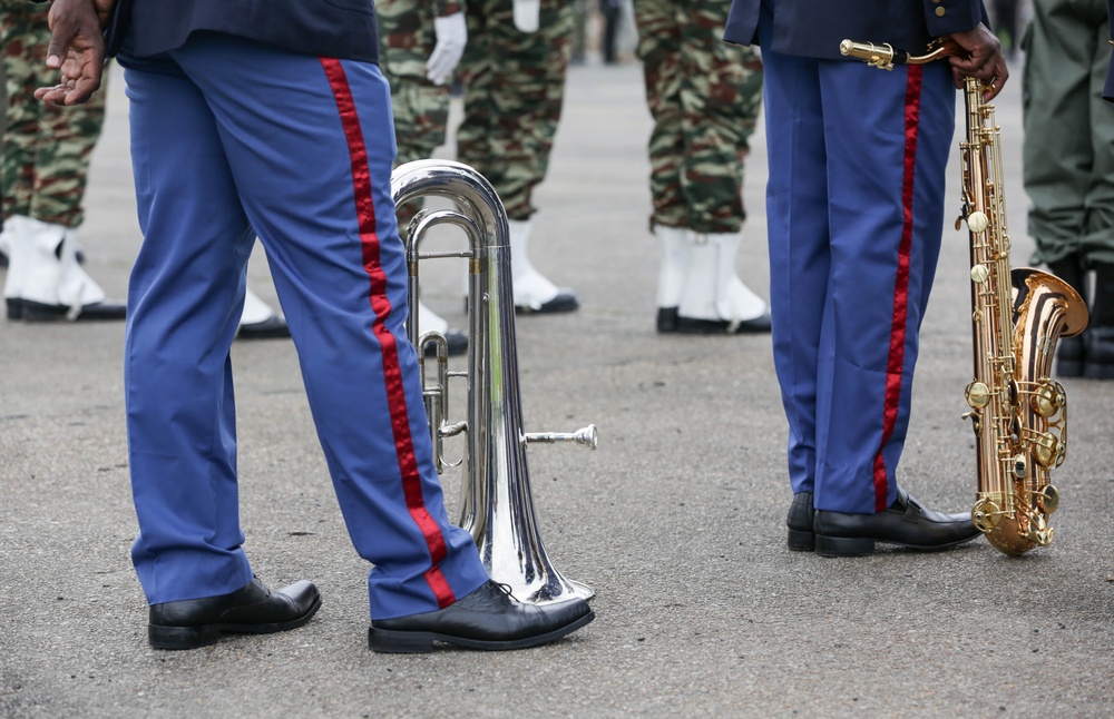 The image shows the lower bodies of military band members in blue dress uniforms with red stripe trim, standing on a paved surface. Two band members are holding musical instruments — a silver euphonium/baritone horn and a gold saxophone. In the background, additional personnel in camouflage uniforms are visible, suggesting a formal military parade or ceremony.